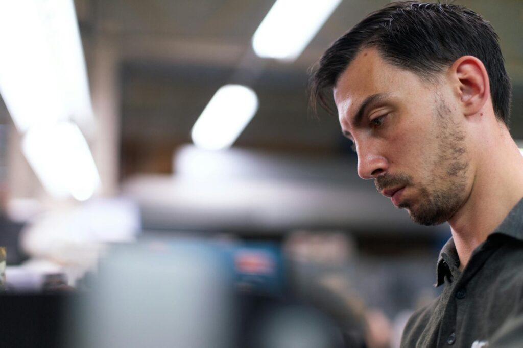 A man in a workshop focuses intently, surrounded by blurred tools and equipment.